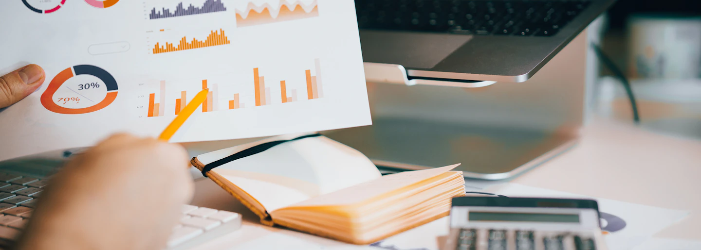 A printed report of coloured charts held above a desk with a notebook and calculator