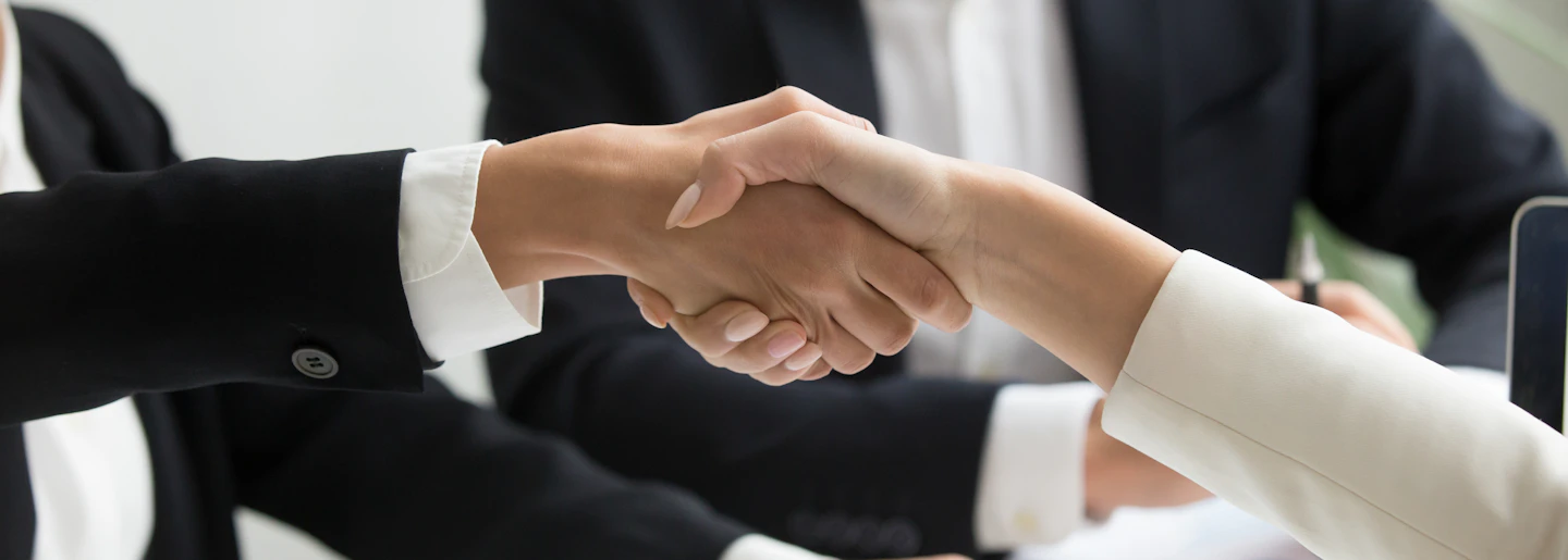 Two people in business dress shaking hands over documents on a desk