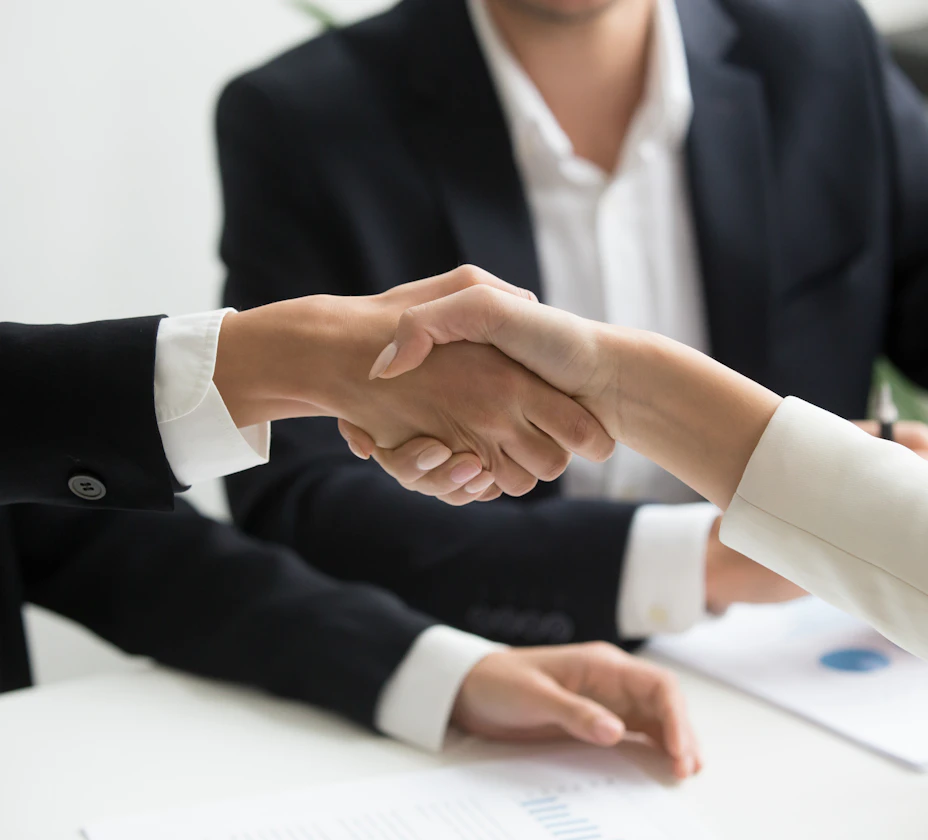 Two people in business dress shaking hands over documents on a desk