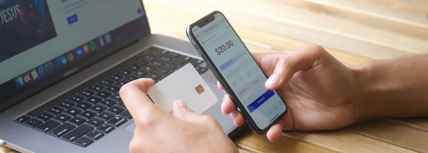 Hands holding a phone showing a banking app while holding a bank card, with a laptop behind