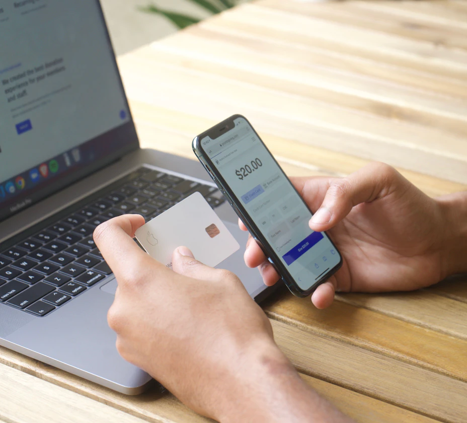 Hands holding a phone showing a banking app while holding a bank card, with a laptop behind